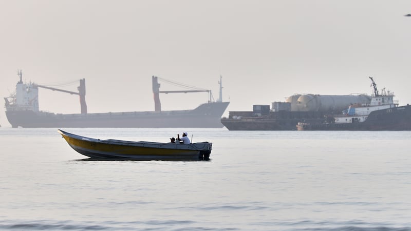 A small boat moves along the shoreline where an a cargo vessel, tugboat and industrial barge...
